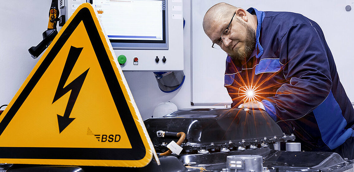 Leadec employee checks a defective battery, in the foreground a warning sign for working under high voltage.