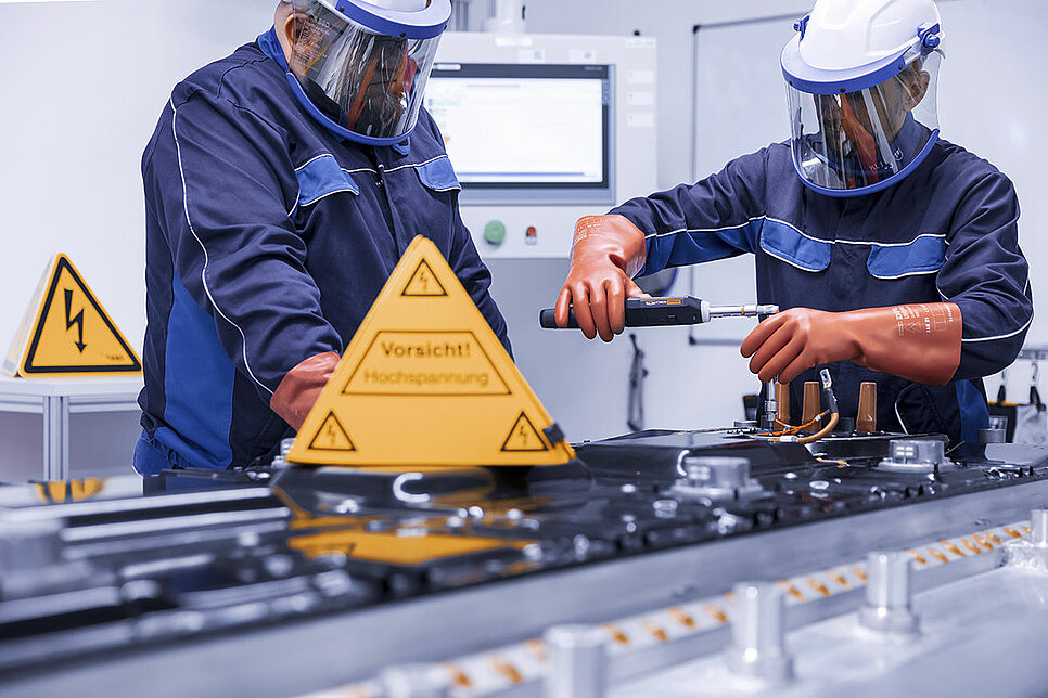 Zwei Leadec-Mitarbeiter in Schutzkleidung bei der Reparatur einer defekten Batterie, vor ihnen ein Warnschild für Arbeiten unter Hochspannung.
