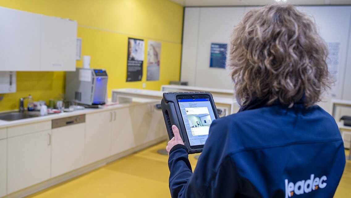 Person with a tablet and a Leadec jacket scans a room. 