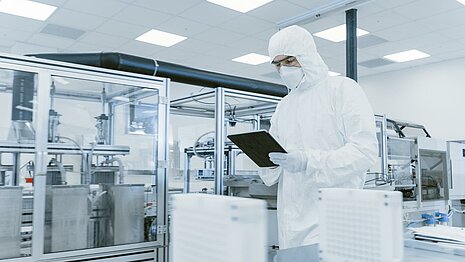 Man in protective suit, mask and goggles looking at a tablet in a semiconductor production facility.