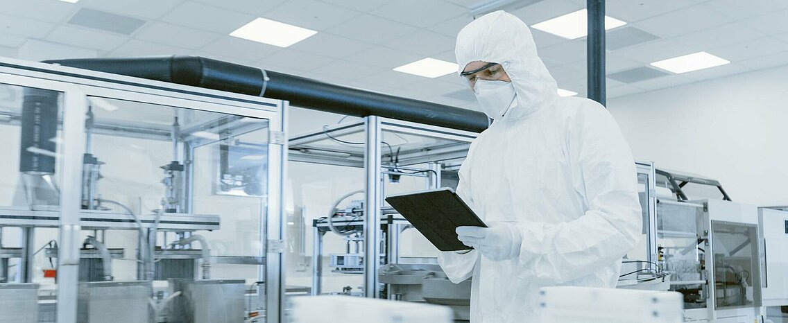 Man in protective suit, mask and goggles looking at a tablet in a semiconductor production facility.