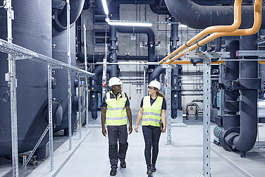 Two Leadec employee wearing safety vests and helmets walk through a technical area.