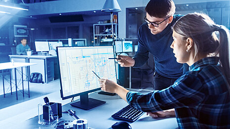 Two people are sitting in front of a screen and are working on the planning of a factory. 
