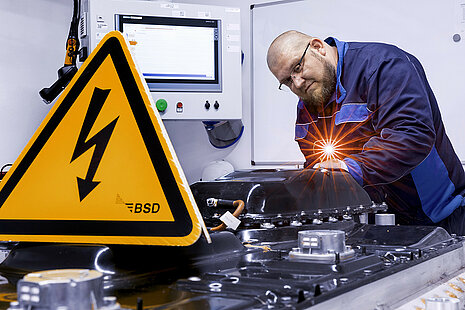 Leadec employee checks a defective battery, in the foreground a warning sign for working under high voltage.