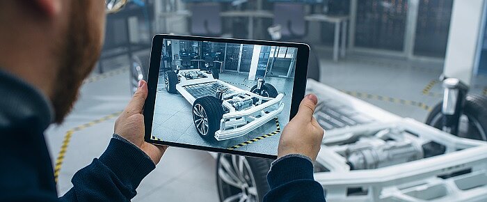 Person mit Tablet in der Hand, im Hintergrund das Grundgerüst eines Elektrofahrzeugs. 