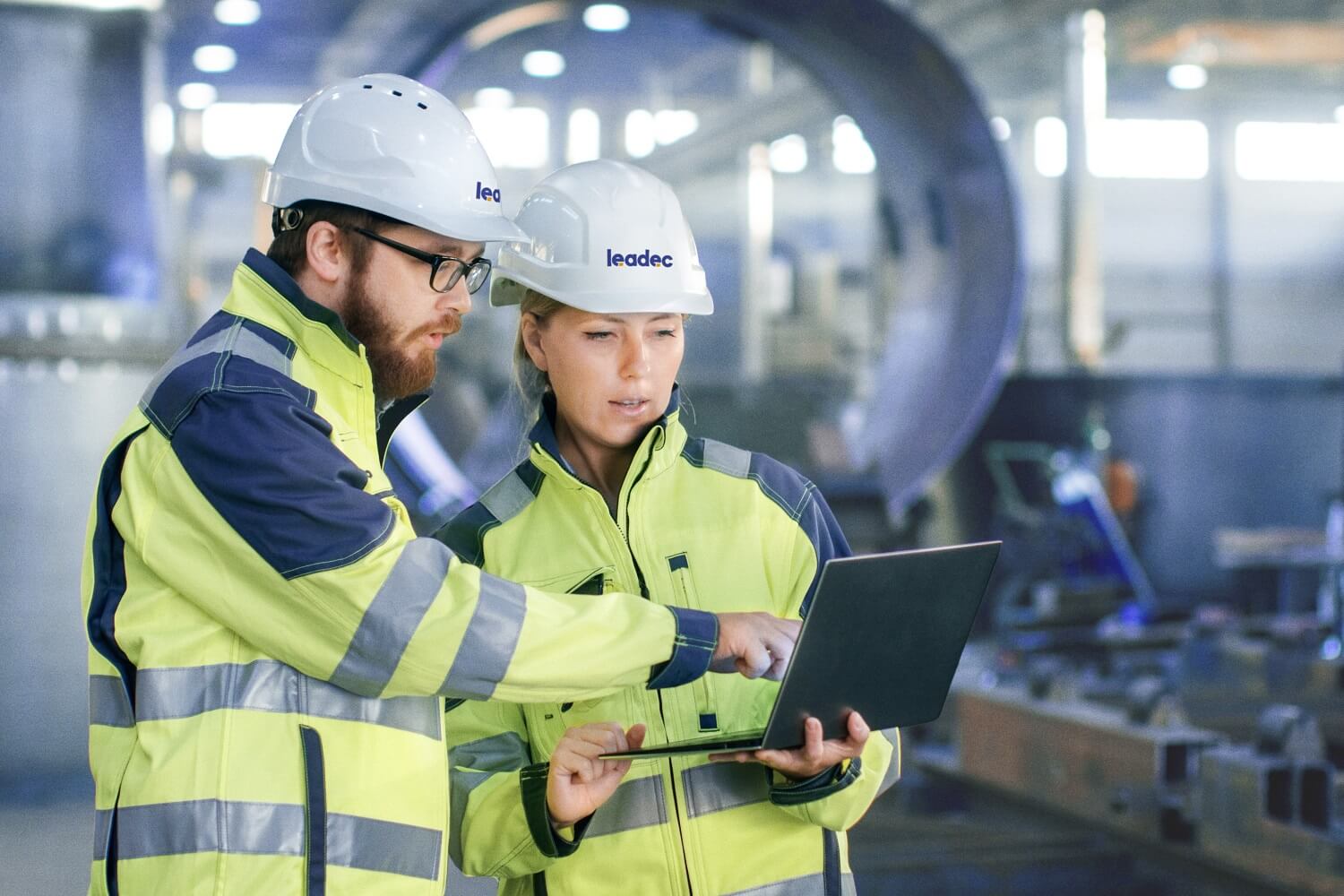 Man and woman inside a factory, both wearing safety clothing and helmets.