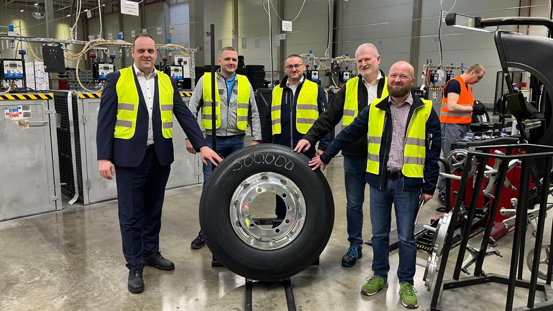 Five men in a factory standing behind a big truck wheel