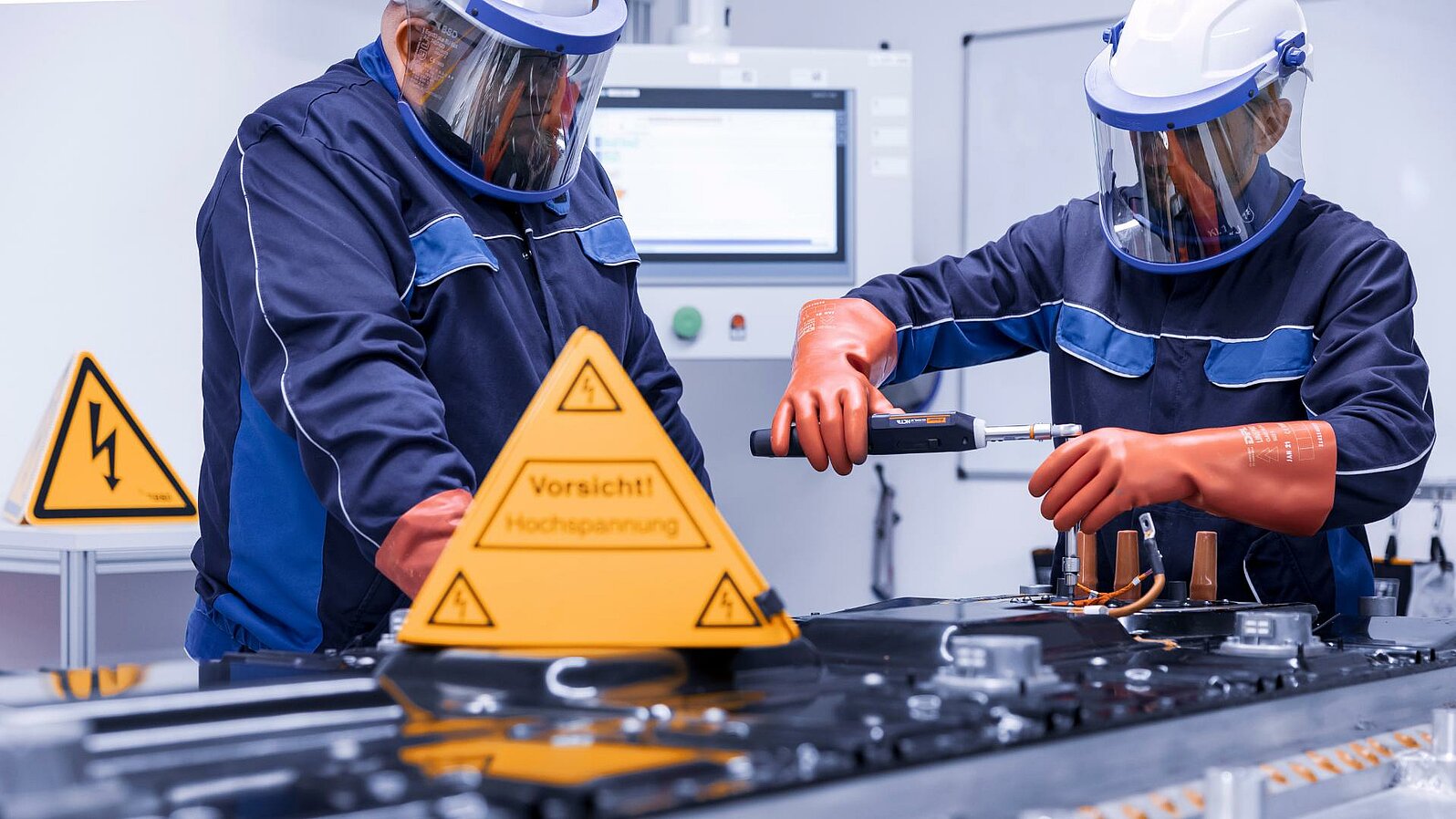 Zwei Leadec-Mitarbeiter in Schutzkleidung bei der Reparatur einer defekten Batterie, vor ihnen ein Warnschild für Arbeiten unter Hochspannung.
